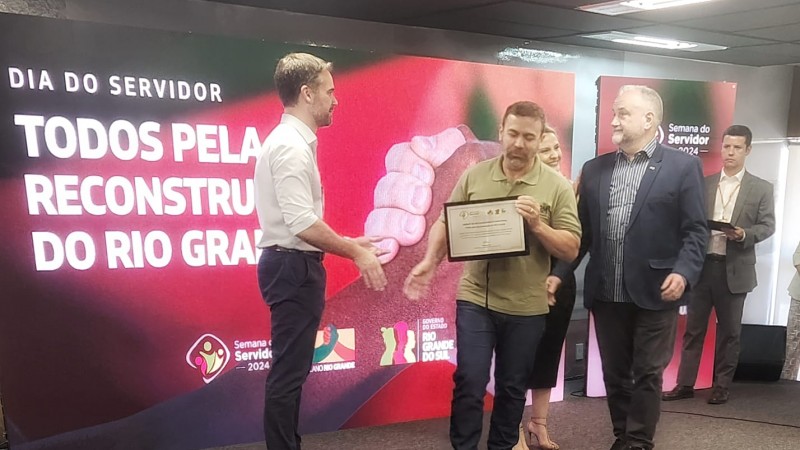 Na imagem, o Governador Eduardo Leite entrega uma placa de homenagem a Luis Adelcio, funcionário da CRM (Companhia Riograndense de Mineração), reconhecido por sua atuação. Luis Adelcio, posicionado no centro, recebe a placa com as duas mãos, enquanto sorri ao lado do Governador, à sua esquerda, e do Presidente Ademir Baretta, à sua direita. Os três são bem humorados, com sorrisos expressivos, em um ambiente formal e iluminado. A placa de homenagem, segurada por Luis Adelcio, representa o reconhecimento pelo trabalho de sua equipe no enfrentamento de desafios recentes. Todos vestem trajes formais, simbolizando a importância da ocasião