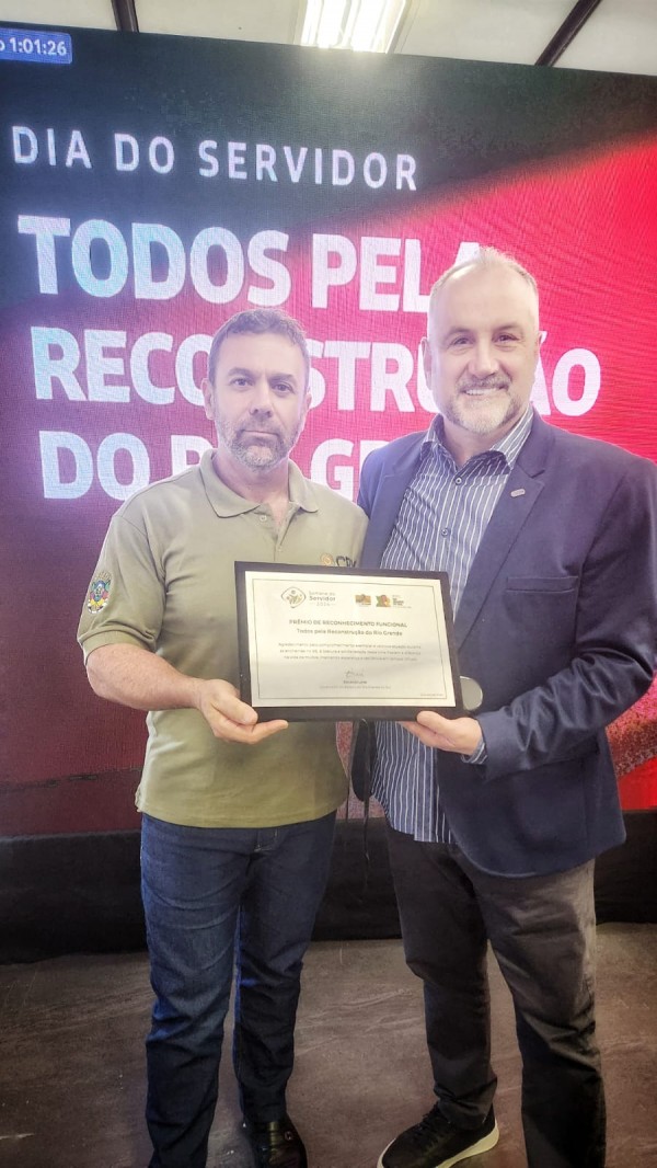 
A imagem mostra o Presidente Ademir Baretta ao lado do funcionário Luis Adelcio. Ambos sorrindo e seguram juntos uma placa de homenagem, com os olhares voltados para a câmera. Luis Adelcio, que recebeu essa homenagem após uma votação online, representa a subsecretaria ou departamento reconhecido pelo importante papel desempenhado no enfrentamento dos efeitos do desastre meteorológico causado pelas enchentes. A placa contém detalhes do prêmio e o nome do órgão, destacando o esforço conjunto da equipe. Ambos os homens vestem roupas formais, com a expressão de satisfação.