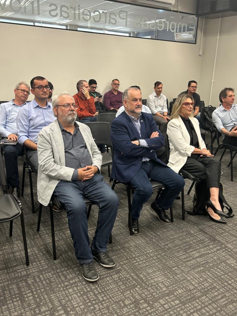 Na imagem mostra na primeira fila 3 pessoas sentadas lado a lado em uma sala de reunião durante o encontro de final de ano da Associação Brasileira do Carbono Sustentável (ABCS). À esquerda, o Diretor João Batista da CRM, de expressão mais séria, veste tons mais escuro e olha para frente. Ao centro, o presidente da CRM, Ademir Baretta, também trajando Blaiser e calça jeans, observa atentamente a reunião. À direita, Asdrid Barato, presidente do Sindicato da Indústria de Extração de Carvão de Santa Catarina, está igualmente vestida de forma mais informal, mantendo uma postura receptiva. O ambiente é profissional, discreto e elementos típicos de uma sala de conferências