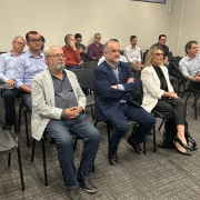 Na imagem mostra na primeira fila 3 pessoas sentadas lado a lado em uma sala de reunião durante o encontro de final de ano da Associação Brasileira do Carbono Sustentável (ABCS). À esquerda, o Diretor João Batista da CRM, de expressão mais séria, veste tons mais escuro e olha para frente. Ao centro, o presidente da CRM, Ademir Baretta, também trajando Blaiser e calça jeans, observa atentamente a reunião. À direita, Asdrid Barato, presidente do Sindicato da Indústria de Extração de Carvão de Santa Catarina, está igualmente vestida de forma mais informal, mantendo uma postura receptiva. O ambiente é profissional, discreto e elementos típicos de uma sala de conferências