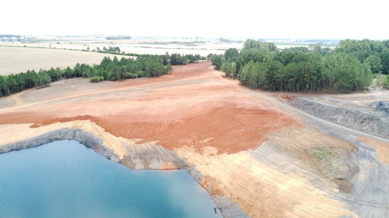 A imagem retrata uma área que passou por recuperação ambiental após a extração de carvão mineral em Iruí, no município de Cachoeira do Sul. No primeiro plano, observa-se um terreno que, anteriormente degradado, agora apresenta vegetação nativa em processo de crescimento. As plantas são de diferentes tamanhos e espécies, criando um ambiente mais diversificado e saudável. Ao fundo, é possível ver uma colina coberta por árvores, evidenciando a recuperação da paisagem. O céu está limpo e ensolarado, simbolizando esperança e renovação. A cena destaca os esforços de restauração ambiental e a importância da preservação dos ecossistemas.