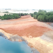 A imagem retrata uma área que passou por recuperação ambiental após a extração de carvão mineral em Iruí, no município de Cachoeira do Sul. No primeiro plano, observa-se um terreno que, anteriormente degradado, agora apresenta vegetação nativa em processo de crescimento. As plantas são de diferentes tamanhos e espécies, criando um ambiente mais diversificado e saudável. Ao fundo, é possível ver uma colina coberta por árvores, evidenciando a recuperação da paisagem. O céu está limpo e ensolarado, simbolizando esperança e renovação. A cena destaca os esforços de restauração ambiental e a importância da preservação dos ecossistemas.