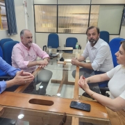 A imagem retrata a reunião do presidente da Companhia Riograndense de Mineração (CRM), Ademir Baretta, com importantes lideranças municipais da metade Sul do Estado. Todos sentados, conversando. À sua esquerda, está o prefeito de Pinheiro Machado, Ronaldo Madruga, que é acompanhado pela vice-prefeita, Laura Ratto Finkler, à sua frente. À direita dele Baretta, encontra-se o prefeito de Candiota, Luiz Carlos Folador á sua frente. Todos estão em um ambiente de reunião, demonstrando um clima de colaboração e diálogo. As expressões dos participantes refletem seriedade e comprometimento com os assuntos discutidos.