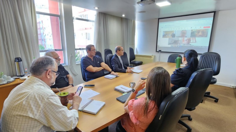 Imagem de uma reunião do conselho de administração em ambiente corporativo. Alguns participantes estão presencialmente ao redor de uma grande mesa de reuniões, enquanto outros aparecem em uma tela, participando remotamente. No centro da mesa, documentos e anotações indicam a pauta discutida. Entre os presentes, há membros do conselho e parte da equipe jurídica, que acompanham atentamente a discussão. O clima é formal, mas positivo, refletindo a comunicação de um acordo favorável para a CRM.