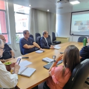 Imagem de uma reunião do conselho de administração em ambiente corporativo. Alguns participantes estão presencialmente ao redor de uma grande mesa de reuniões, enquanto outros aparecem em uma tela, participando remotamente. No centro da mesa, documentos e anotações indicam a pauta discutida. Entre os presentes, há membros do conselho e parte da equipe jurídica, que acompanham atentamente a discussão. O clima é formal, mas positivo, refletindo a comunicação de um acordo favorável para a CRM.