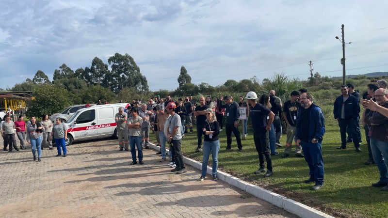 imulado de evacuação de emergência realizado em via pública na cidade de Candiota. A imagem mostra moradores e equipes de resposta caminhando pela rua, seguindo orientações de agentes uniformizados. Algumas pessoas carregam mochilas ou pertences, representando uma evacuação realista. Há viaturas e cones de sinalização no local, e o clima é de organização e atenção. O exercício ocorre durante o dia, com boa visibilidade.