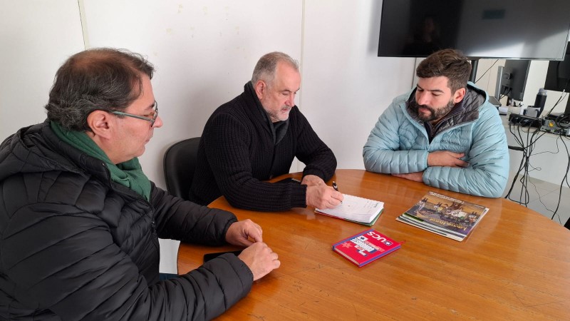 Três pessoas estão reunidas em uma sala de reunião, sentadas ao redor de uma mesa. Na mesa, há alguns papéis espalhados. A cena transmite um ambiente de colaboração e discussão.