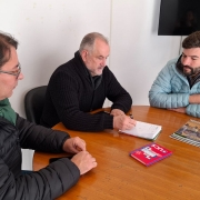 Três pessoas estão reunidas em uma sala de reunião, sentadas ao redor de uma mesa. Na mesa, há alguns papéis espalhados. A cena transmite um ambiente de colaboração e discussão.