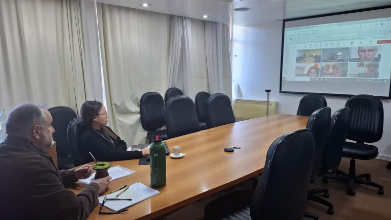 Na imagem, há duas pessoas presenciais em uma sala de reunião, ambas com expressão séria e concentrada. No online, as expressões são sérias e dedicadas à reunião. A cena transmite um ambiente de trabalho focado e colaborativo em um formato híbrido