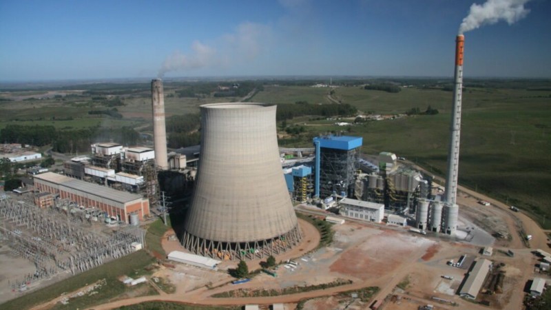 Imagem de uma usina termel&eacute;trica localizada em Candiota. No centro, aparecem grandes estruturas industriais feitas de concreto e metal, com torres altas e chamin&eacute;s emitindo colunas de fuma&ccedil;a clara. Ao redor, h&aacute; tubula&ccedil;&otilde;es, tanques e pr&eacute;dios operacionais. No primeiro plano, v&ecirc;-se uma &aacute;rea aberta com solo escuro, t&iacute;pico da regi&atilde;o de extra&ccedil;&atilde;o de carv&atilde;o mineral. O c&eacute;u est&aacute; parcialmente nublado, destacando o contraste entre as estruturas da usina e o ambiente ao redor.