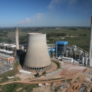 Imagem de uma usina termel&eacute;trica localizada em Candiota. No centro, aparecem grandes estruturas industriais feitas de concreto e metal, com torres altas e chamin&eacute;s emitindo colunas de fuma&ccedil;a clara. Ao redor, h&aacute; tubula&ccedil;&otilde;es, tanques e pr&eacute;dios operacionais. No primeiro plano, v&ecirc;-se uma &aacute;rea aberta com solo escuro, t&iacute;pico da regi&atilde;o de extra&ccedil;&atilde;o de carv&atilde;o mineral. O c&eacute;u est&aacute; parcialmente nublado, destacando o contraste entre as estruturas da usina e o ambiente ao redor.