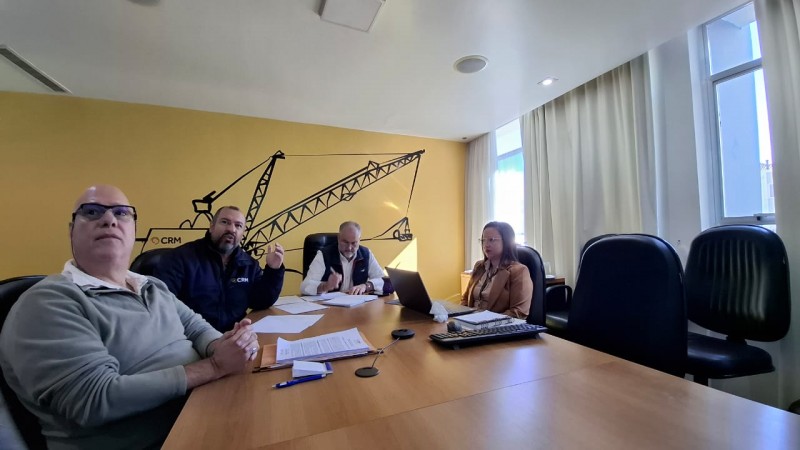 4 pessoas est&atilde;o numa sala ampla de olhos na tela, porque est&aacute; acontecendo a reuni&atilde;o de administra&ccedil;&atilde;o mensal. No momento apenas 1 est&aacute; falando e todos est&atilde;o prestando aten&ccedil;&atilde;o. A mesa &eacute; retangular e aparecem os 4 que est&atilde;o na sala da CRM