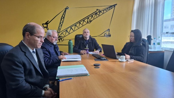 Numa sala ampla quatro pessoas est&atilde;o sentadas numa reuni&atilde;o de olho na tela onde est&aacute; acontecendo a reuni&atilde;o online, rodos atentos no que est&aacute; acontecendo. 