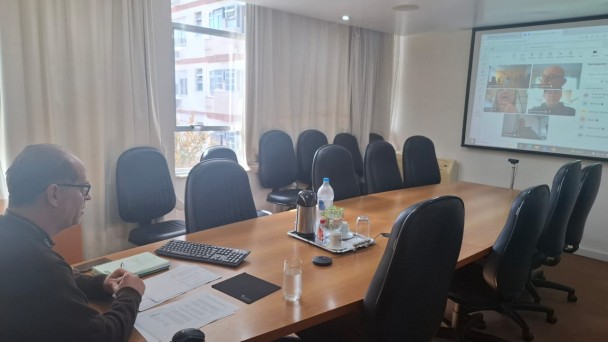 Uma pessoa ao centro da mesa em reuni&atilde;o virtual com os demais participantes que est&atilde;o online e na tela. todos est&atilde;o atentos &agrave; reuni&atilde;o. 